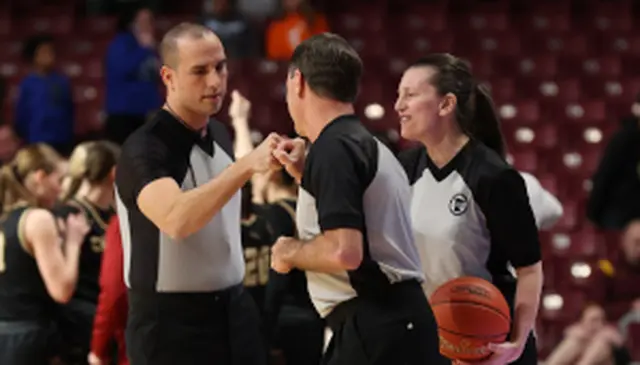 Three basketball referees in black and white uniforms fist-bump on the court, with one holding a basketball.