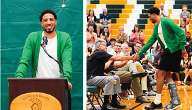 A man in a green cardigan speaks at a podium and shakes hands with a seated man in a gymnasium with a cheering crowd.